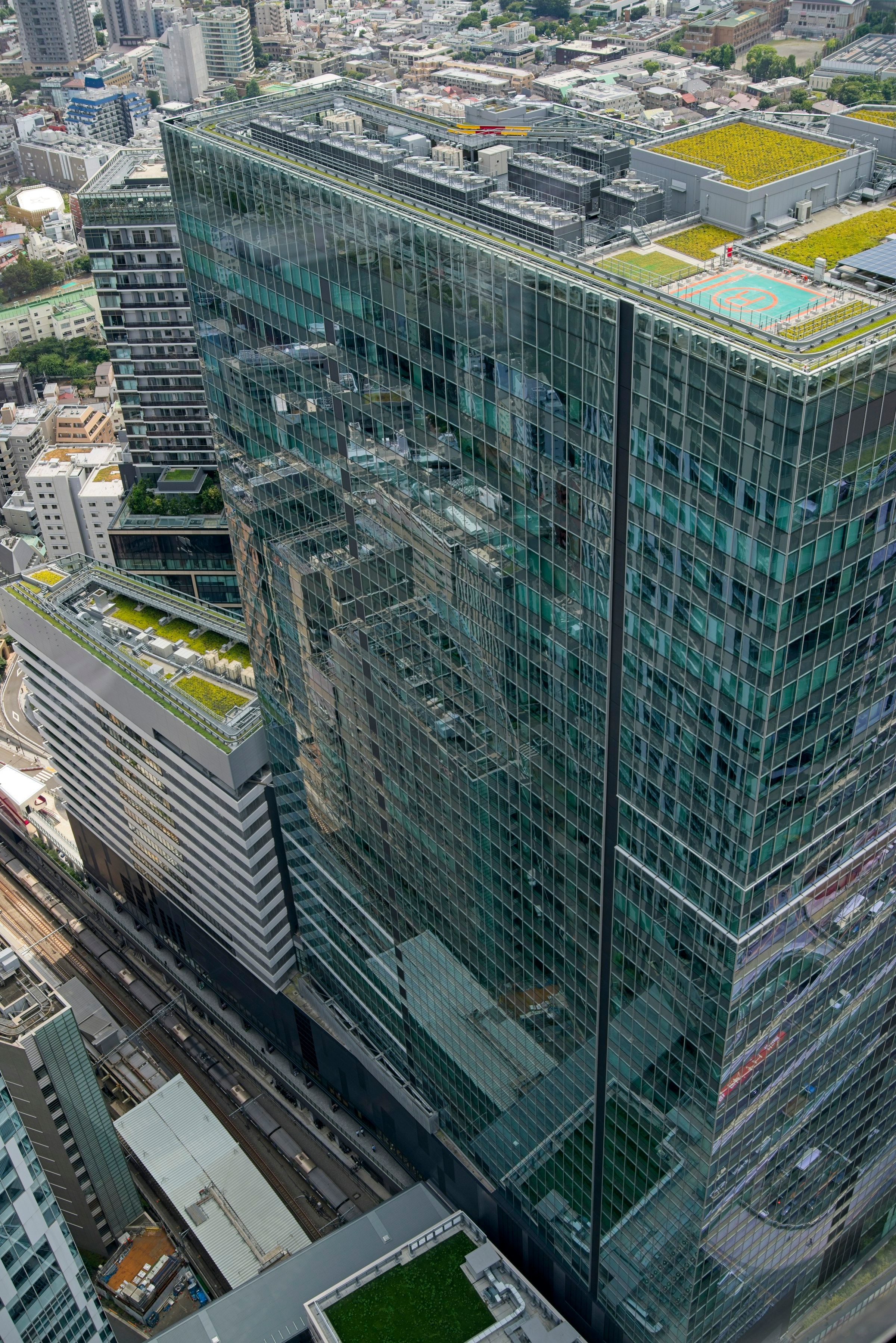 An aerial view of a green-glass skyscraper whose rooftop holds a helipad, with a dense city behind it.