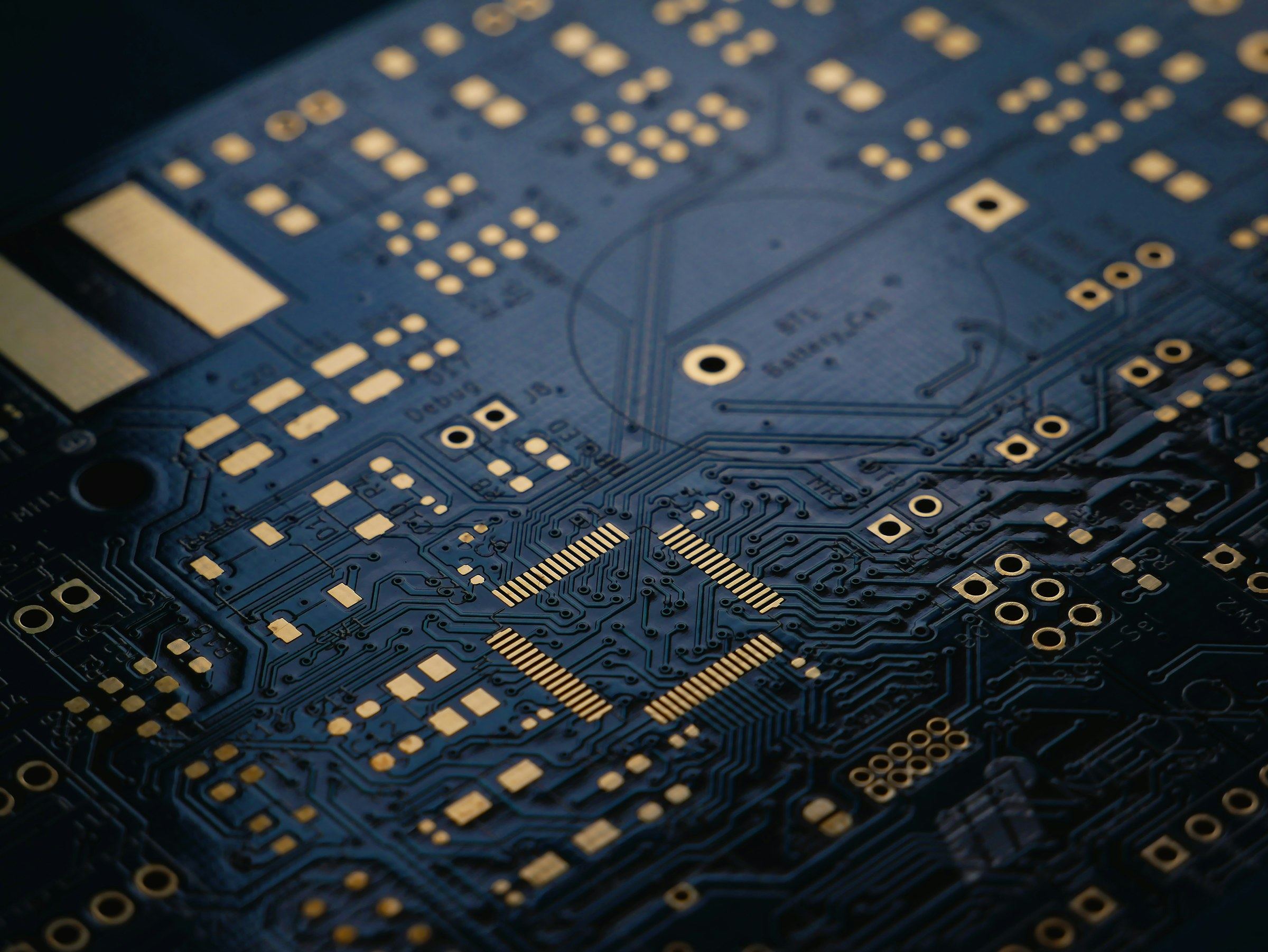 Close-up of a dark blue printed circuit board with gold solder pads and fine traces receding into soft focus.